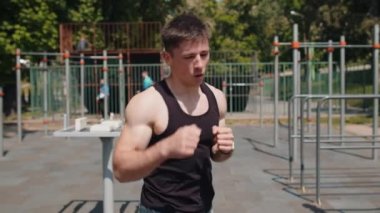 Spor giyim kuşağında beyaz atletik kaslı bir adam gölge boksu aerobik kardiyografisi eğitimi alıyor. Çocuk parkında tişörtlü genç bir adam. Rutin çalışma. Güç ve motivasyon. Açık hava spor salonu