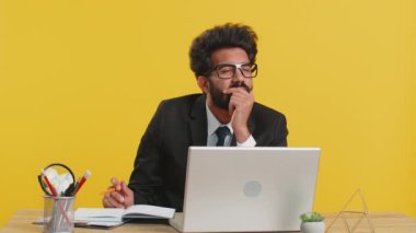 Düşünceli Hintli gazeteci iş adamı notlar alıyor, not defterine kalemle düşünceler yazıyor, liste yapıyor, ofis masasında iyi bir fikir. Dizüstü bilgisayarda çalışan takım elbiseli Hintli adam.