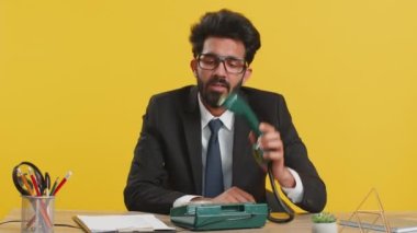 Hintli işadamı, 80 'lerin telli vintage telefonunda can sıkıcı konuşmalar yaparak ofis masasında aptal suratlar yaparak aptal durumuna düşüyor. Sarı arka planda izole edilmiş resmi takım elbiseli Hindu adam.