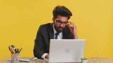 Ofis masasında kameraya bakıp gözlüğünü çıkaran mutlu, sakallı Hintli işadamının portresi. Resmi takım elbiseli bir erkek Hindu. Yakışıklı uzaktan kumandalı adam dizüstü bilgisayarla çalışıyor