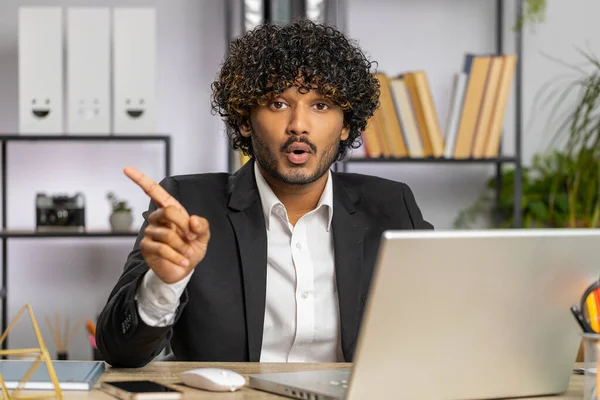 Indian young businessman working on laptop computer shakes finger and saying No be careful scolding and giving advice to avoid danger mistake disapproval sign at home office. Confident freelancer man
