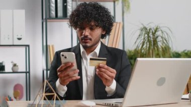 Sakallı iş adamı yazılım geliştirici kredi kartı ile online satın alma alışverişi yapıyor ve ofis masasında dizüstü bilgisayar kullanıyor. Hintli serbest çalışan iş adamı