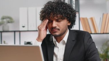 Tired ill indian businessman suffering from headache problem tension and migraine, stress at home office desk workplace. Bearded freelancer broker worker man works on laptop notebook. Overworking