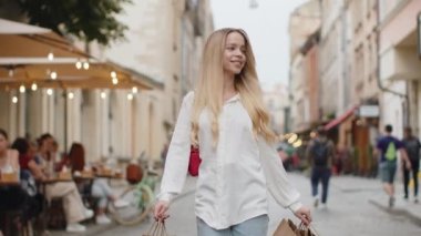 Açık havada hediyelerle dolu poşetlerle alışveriş yaptıktan sonra gülümseyen sarışın genç kadın tüketici. Şehir caddesi boyunca yürüyen genç turist kız gezgini. Kasaba yaşam tarzları