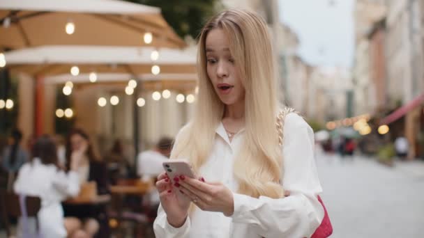 ブロンドの若い女性は モバイルスマートフォンを使用しています 都会の街を歩く幸せなティーンエイジャーの女の子の観光客 町の暮らし — ストック動画