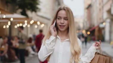 Alışveriş sonrası alışveriş meraklısı mutlu bir genç kadın. Açık havada hediye dolu torbalarla. Genç kız turist şehir caddesinde yürüyüp akıllı telefon konuşmaları yapıyor.