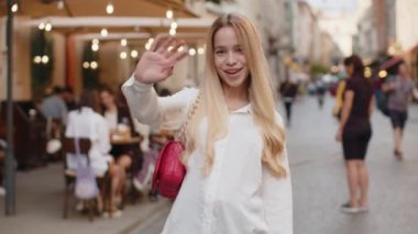 Sarışın genç bayan kameraya dostça gülümsüyor, el sallıyor merhaba, selam veriyor ya da veda ediyor, misafirperver bir yüz ifadesiyle karşılıyor. Şehir caddesinde yürüyen genç kız.