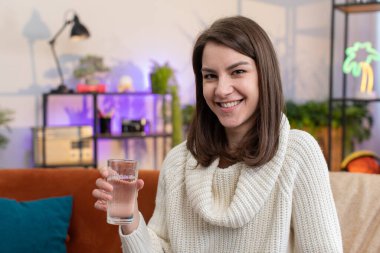 Thirsty young woman holding glass of natural aqua make sips drinking still water preventing dehydration sits at home living room. Girl with good life habits, healthy slimming, weight loss concept