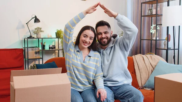 Young family marriage couple man woman raise hands join arms together making house roof shape, symbol of new home, happy homeowners, rental flat, bank loan, insurance. Husband wife on sofa in room