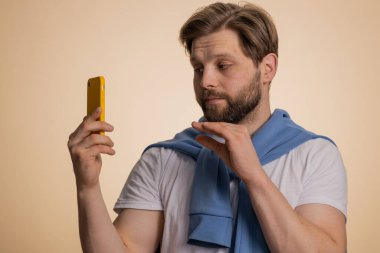 İlgisiz beyaz bir adam akıllı telefonda can sıkıcı konuşmalar yapıyor. Uykulu yetişkin adam arkadaşıyla cep telefonuyla konuşuyor, çevrimiçi konuşmaları stüdyo bej arka planında izole ediyor.