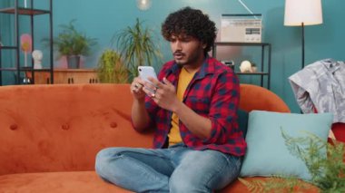 Üzgün Hintli adam akıllı telefon daktilo kullanıyor. Ani piyango sonuçlarını kaybediyor. Kötü haber, servet kaybı, başarısızlık, teslim tarihi, virüs. Evdeki kanepede oturan Hintli genç adam.