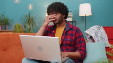 Tired freelancer indian man use laptop, suffering from headache problem tension and migraine, stress at home room. Hindu guy works on notebook, sends messages, makes online purchases, watching movies