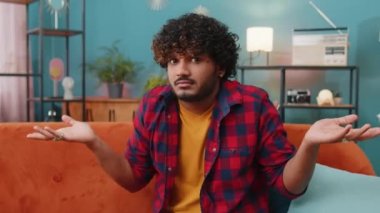 I dont know what to say. Confused indian handsome man feeling embarrassed about ambiguous question, having doubts, no idea, being clueless and uncertain at home. Hindu guy sits on sofa in living room