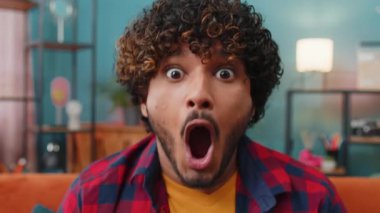 Aman Tanrım, vay canına. Genç Hintli adam kameraya büyük gözlerle bakarken şaşırdı. Ani bir zafer, iyi bir galibiyet haberi, kutlama. Evdeki kanepede oturan heyecanlı Hintli adamın portresi.