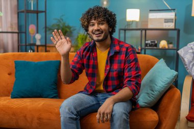 Hello. Indian handsome man smiling friendly at camera and waving hands gesturing hello, hi, greeting or goodbye, welcoming with hospitable expression at home. Hindu guy sitting on sofa in living room