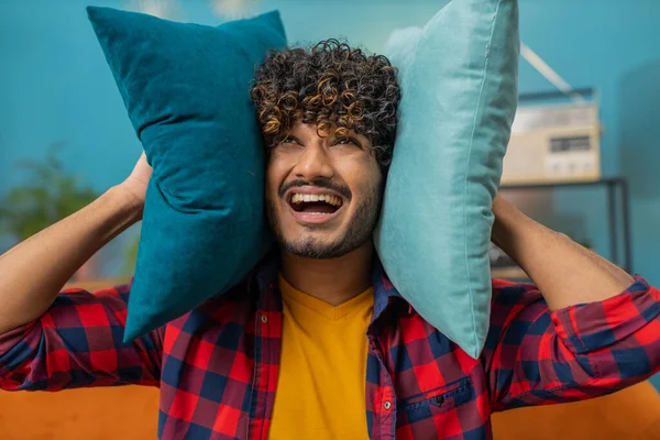Repair work at neighbours. Irritated indian man relaxing on couch cover ears with pillows annoyed by noisy neighbors suffer from headache wish silence. Thin walls at home flat without sound insulation
