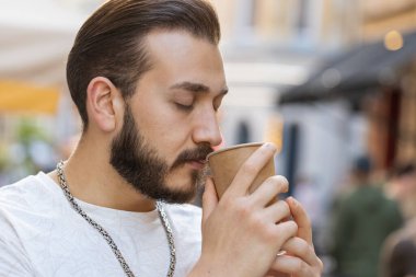 Mutlu sakallı delikanlıya yakın çekim sabah kahvesinin tadını çıkarıyor ve dışarıda gülümsüyor. Dinleniyorum, ara veriyorum. Şehirdeki Sunshine Caddesi 'nde yürüyen ve kahve içen bir adam. Kasaba yaşam tarzları