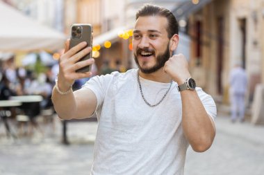 Sakallı genç adam cep telefonuyla kutlama yapıyor. İyi haber kazan, piyango zaferi kazan, açık havada dağıtım yap. Şehir Güneşi Şehri caddesinde yürüyen mutlu bir turist. Kasaba yaşam tarzları