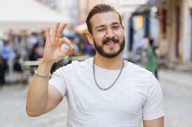 - Tamam. Mutlu, neşeli genç adam kameraya onaylayıcı bir şekilde bakıyor. Tamam jestini gösteriyor. İşaret gibi olumlu bir şey. Neşeli adam şehir güneşli caddede yürüyor, açık havada.