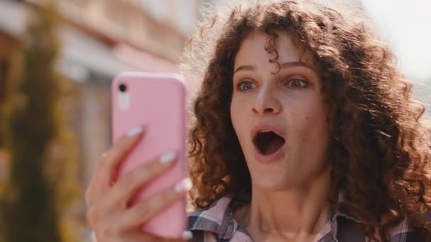 うわぁ 喜びに満ちた幸せな勝者かなり若い女性は良いメッセージニュースを受賞を祝うスマートフォンのタイピングブラウジングを使用しています 携帯電話を持つ女の子は屋外の都会の日差しの街で歩く — ストック動画