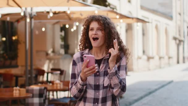 かなり若い女性は 良いメッセージニュース 宝くじのジャックポットの勝利 プレゼントをオンラインで屋外で受賞を祝う携帯電話のスマートフォンを使用しています 都会の日差しの街を歩く幸せな女の子の観光客 町の暮らし — ストック動画