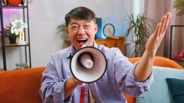 Asian handsome man talking with megaphone, proclaiming news, loudly announcing advertisement, warning using loudspeaker to shout speech, real estate sale at home. Chinese guy sitting on sofa in room