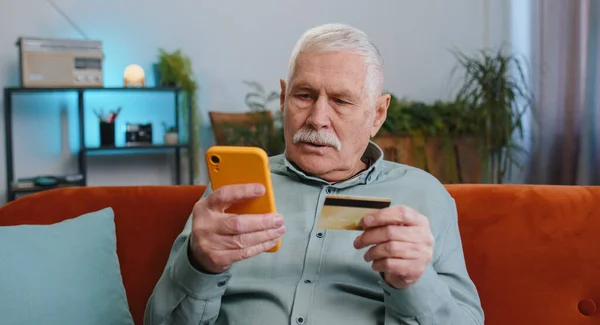Grandfather senior man using credit bank card and smartphone while transferring money, purchases online shopping cashless, order food delivery, pay bills at home apartment indoor. Elderly guy on couch