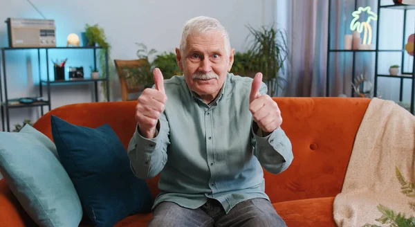 快乐兴奋的老爷爷赞许地看着摄像机露出大拇指 就像签了字一样肯定的东西 好消息 积极的反馈 老年人坐在家里房间的沙发上 — 图库照片
