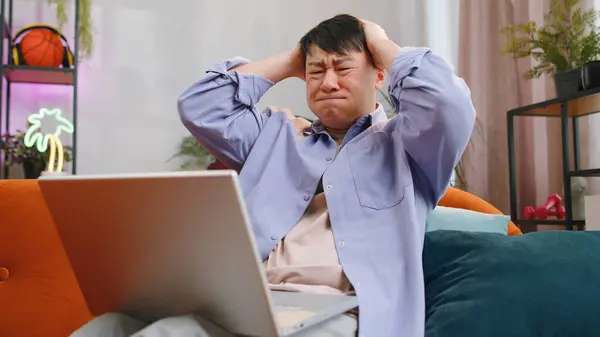 不満アジアの男は ラップトップノートパソコンのノートパソコンのタイプのブラウジング作業を使用して 損失が突然宝くじの結果に驚きになる悪いニュースの幸運損失は コンピュータウイルスに失敗します 中国人男ホームでリビングルームオンソファ — ストック写真