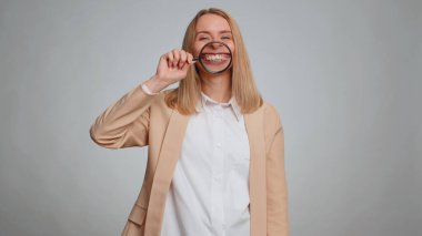 Business woman holding magnifier glass on healthy white teeth, looking at camera with happy expression, showing funny silly face smiling mouth. Secretary office girl isolated on gray studio background
