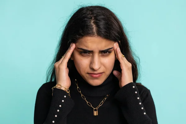 Pretty indian arabian woman in sweater rubbing temples to cure headache problem, suffering from tension, migaine, stress, grimacing in pain, high blood pressure isolated alone on blue wall background