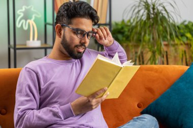 Genç Hintli adam ilginç kitaplar okuyor, sayfaları çevirip gülümsüyor edebiyattan zevk alıyor, rahat bir kanepede dinleniyor. Evdeki kanepede oturan mutlu Hintli adamın portresi.