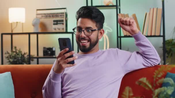 幸せなインドの男は携帯電話のタイプブラウジングを使用すると言うすごいはい勝者のジェスチャーを行う宝くじの大当たりの良いニュースを祝う大きな勝利が判明した ヒンドゥー若いです男ホームで部屋に座っているソファ — ストック動画