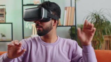 Indian man using virtual reality futuristic technology VR app headset helmet to play simulation 3D 360 video game, watching film movie at modern home apartment. Hindu guy in goggles sitting on couch