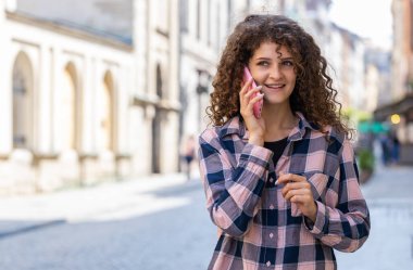 Uzaktan iletişim kuran mutlu genç bayan turist akıllı telefonla konuşuyor. Güzel kız telefonda iyi haberlerin dedikodusunu yapıyor. Şehir caddesinde yürüyor. Şehir yaşam tarzları açık havada