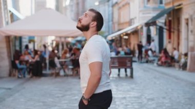 Avrupa caddesinde yürüyen sakallı genç bir turistin kamera karşısına geçip gülümsemesi. Arka planda gezgin bir adamın şehir güneşli şehrinde yürüyüşü var. Kasaba yaşam tarzları