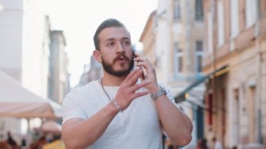 Uzaktan iletişim kuran mutlu genç turist akıllı telefonla konuşuyor. Sakallı adam telefonda iyi haber dedikoduları yapıyor. Şehir caddesinde yürüyor. Şehir yaşam tarzları açık havada