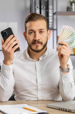 Akıllı telefon gösteren iş adamı içten bir sevinçle kazanıyor, nakit para alıyor, piyango başarısı, telefonla kazanmayı öneriyor. İş yerindeki adam kameraya bakıyor. Dikey