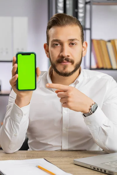Young developer business man hold smartphone with green screen chroma key mock up recommend good application promotional sale offer. Freelancer guy at office workplace looking at camera. Vertical shot