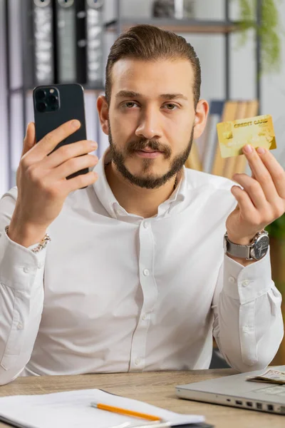 stock image Young middle eastern boss business man using credit bank card and smartphone while transferring money, purchases online shopping ordering delivery. Guy at office workplace looking at camera. Vertical