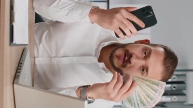 Akıllı telefon gösteren iş adamı içten bir sevinçle kazanıyor, nakit para alıyor, piyango başarısı, telefonla kazanmayı öneriyor. İş yerindeki adam kameraya bakıyor. Dikey