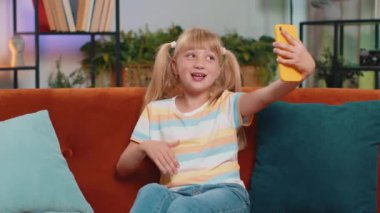 Young blonde child kid girl blogger taking selfie on smartphone, communicating video call online with social media subscribers. Female teen toddler recording video story at home in room sits on sofa