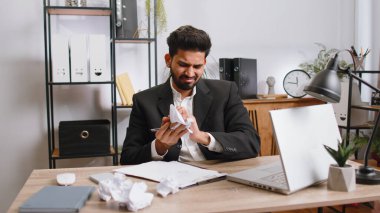 Angry furious indian man working at office throwing crumpled paper, having nervous breakdown at work, migaine, headache, stress management, mental distress problems, losing temper, reaction on failure