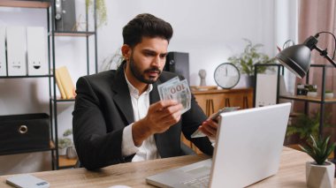 Rich indian businessman working on laptop pc counting money cash, calculate earnings income profit at home office workplace. Professional manager freelancer man. Business people. Employment occupation