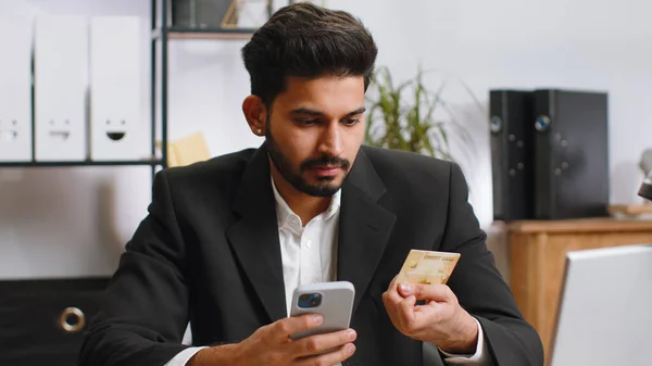 Bearded businessman programmer software developer working making online purchase payment shopping with credit bank card smartphone at home office desk with laptop. Indian freelancer business man