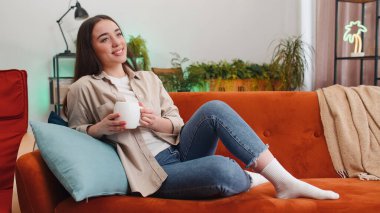 Çekici gülümseyen genç bir kadının portresi bir fincan sıcak kahve ya da kanepede uzanmış bitki çayı içiyor. Oturma odasındaki kız tek başına rahatça dinleniyor.