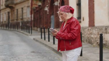 Gezgin son sınıf öğrencisi kadın akıllı telefon kullanıyor açık hava mobil navigatör uygulamasında haritada arama yapmak için kısa mesajlar yazıyor. Yaşlı bir büyükanne şehir sokağının önünden geçiyor.