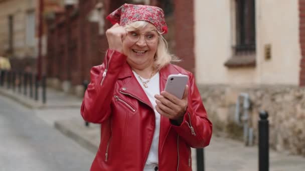 老年妇女使用手机庆祝胜利的消息 彩票中奖 在线赠送 快乐的老奶奶走在城市街道的背景下 生活方式 — 图库视频影像