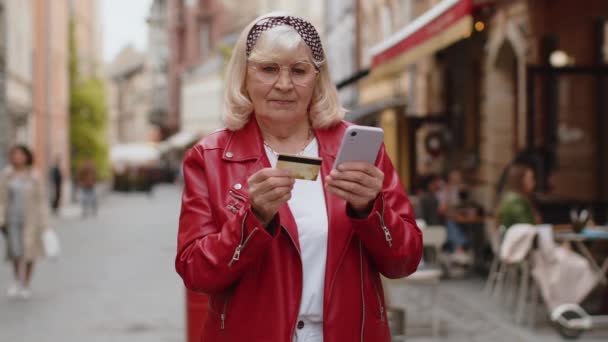 お金を転送しながら クレジットカードのスマートフォンを使用してシニア女性は オンラインショッピングを購入し 食品の配達を注文し ホテルの部屋を予約します 外の都会の通りを歩くおばあちゃんのおばあちゃん — ストック動画