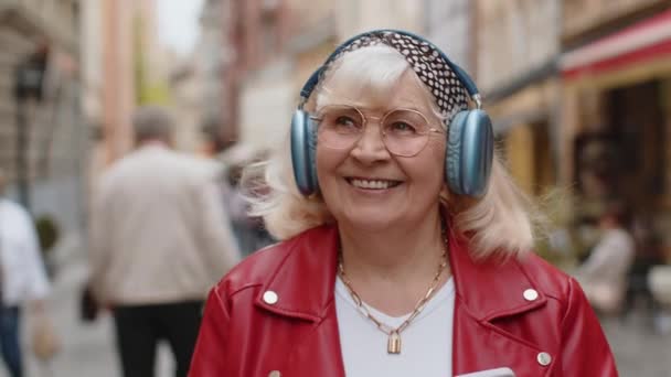 在智能手机户外舞曲中 老年女性在无线耳机中选择听自己喜爱的充满活力的迪斯科摇滚音乐时 开心而轻松的特写镜头让她们喜出望外 奶奶走在城市街道上 — 图库视频影像